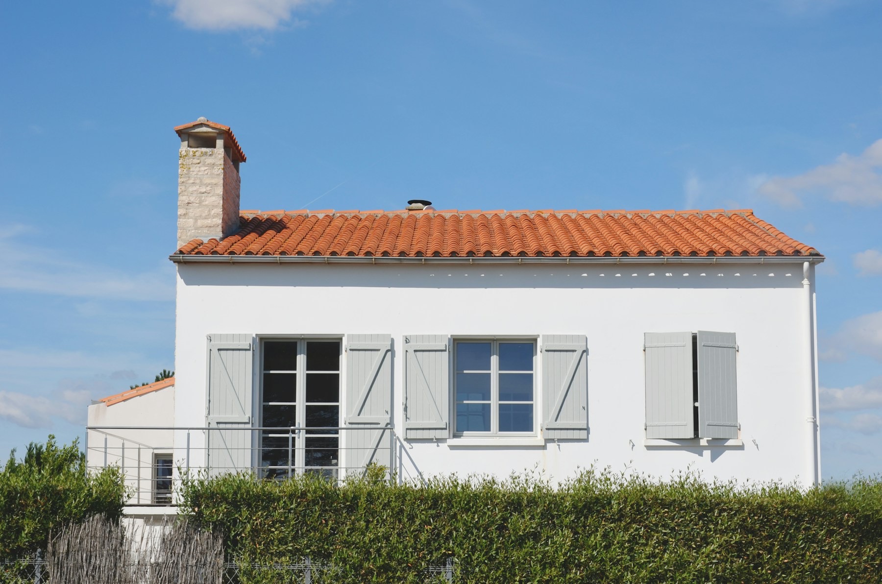 Façade blanche d'une maison à toit de tuiles et volets gris, derrière une haie.